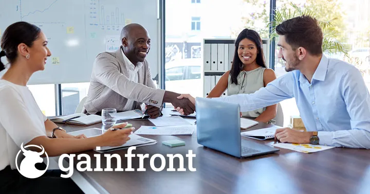Four coworkers in a bright meeting room shaking hands and reviewing paperwork around a laptop, with the Gainfront logo in the corner.