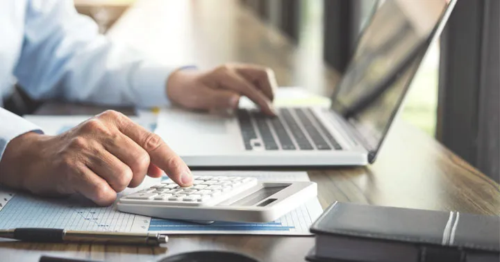 Close-up of a person using a calculator beside an open laptop and paperwork on a desk.